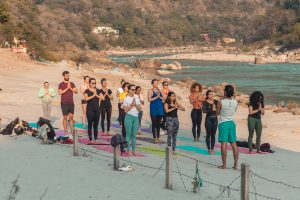 Greeting the sun by the beach: In the tranquil setting of Rishikesh, students at Sage House Yoga engage in beach Acro Yoga, beautifully synchronizing their movements as they perform the Sun Salutation, embracing the energy and warmth of the sun.