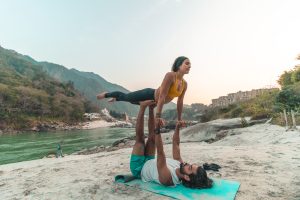 Joyful elevation by the beach: Amidst the serene backdrop of Rishikesh, students at Sage House Yoga revel in the exhilarating practice of beach Acro Yoga, experiencing the thrill and delight of acro lifts with smiles and laughter.