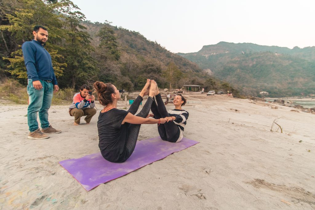 Strength and balance in sync: At the serene beach in Rishikesh, a couple of students gracefully perform the Boat Pose during a beach Acro Yoga session at Sage House Yoga, embodying harmony and core stability.