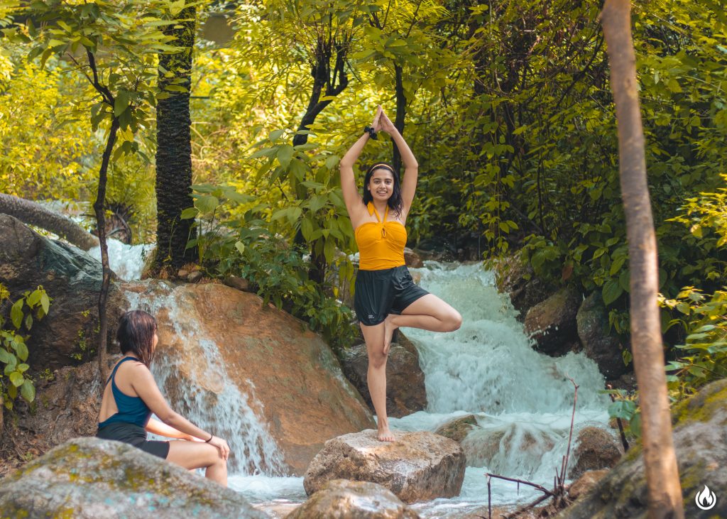Serenity and strength amidst nature: At the Sage House Yoga retreat in Rishikesh, a girl gracefully performs a standing leg pose near a majestic waterfall, finding balance and harmony.
