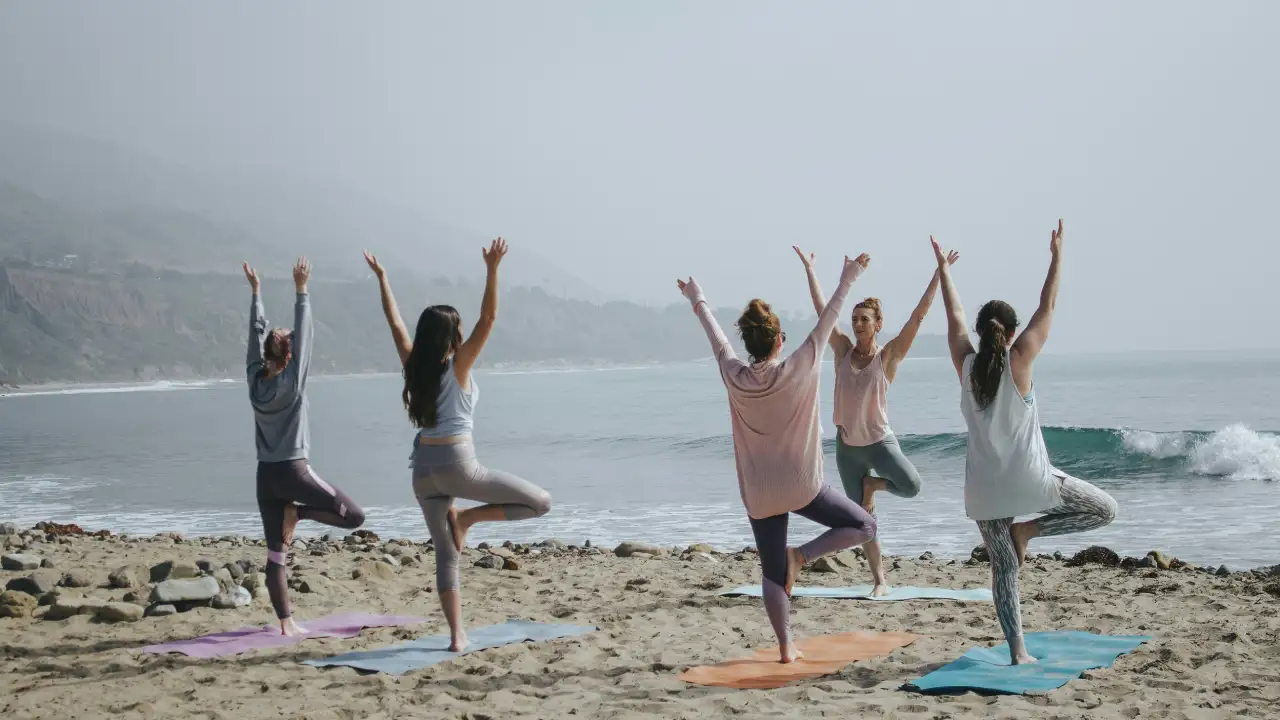 Group Of Women Practicing Yoga In Tree Pose On The Beach | Sage House Yoga In Rishikesh